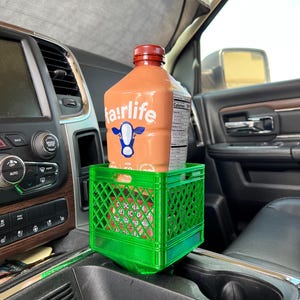 May include: A brown Fairlife chocolate milk bottle with a red cap sits in a small green plastic crate inside a vehicle. The crate is placed in the center console of the car. The Fairlife logo features a blue cow.