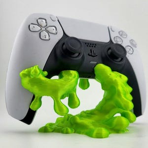 May include: A white and black PlayStation 5 controller with a neon green, 3D-printed slime design. The controller has black joysticks and buttons. The slime appears to be dripping from the bottom of the controller.
