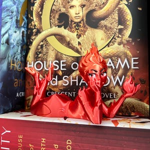 May include: A red, stylized figurine of a woman with flames for hair, resting on a stack of books. The figurine is a bookmark, with the title "House of Flame and Shadow" visible behind it. The figurine is approximately 4 inches long.