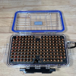 May include: A clear plastic storage case with a blue rubber seal containing a black foam insert filled with rows of copper-jacketed ammunition cartridges. The case features a hinged lid, a black latch, and a small metal carabiner attached to the side.