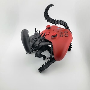 May include: A black and red sculpture of a creature with a red game controller as its back. The creature has a long, segmented tail and spiky details. The controller has red buttons and a smooth surface.