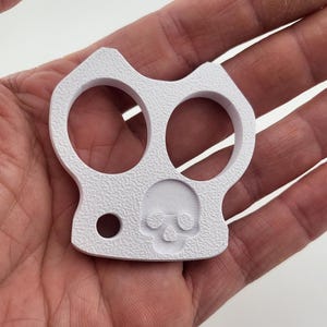 May include: A white, textured knuckle duster with two finger holes and a small hole. A skull is embossed on the front. The object is held in a person's hand.
