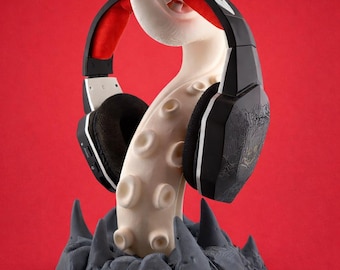 May include: A black and red headset rests on a white octopus tentacle-shaped stand. The stand is mounted on a gray, rocky base, all against a red background. The headset has black earcups and a red headband.