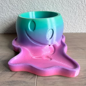 May include: A pink and green ombre ghost-shaped planter with a smiley face. The planter has a round top and a dripping base.