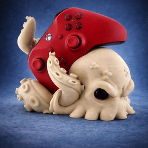 May include: A red video game controller rests atop a cream-colored octopus sculpture. The controller has black buttons and a white logo. The octopus has large, dark eyes and detailed tentacles. The background is a gradient of blue.