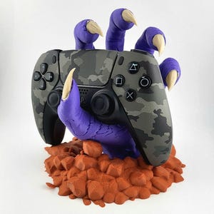 May include: A camouflage-patterned video game controller held by a purple claw with tan nails, resting on an orange, rocky base. The controller has black buttons and directional pads. A unique gaming accessory.