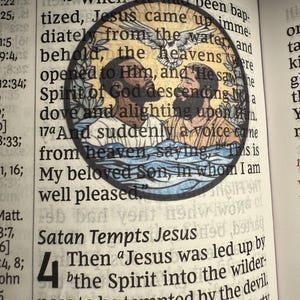 Puede incluir: Ilustraci&oacute;n circular de vidriera que representa una escena religiosa con dos figuras en el agua, una paloma y texto de la Biblia. La imagen est&aacute; situada sobre un libro abierto con texto impreso.