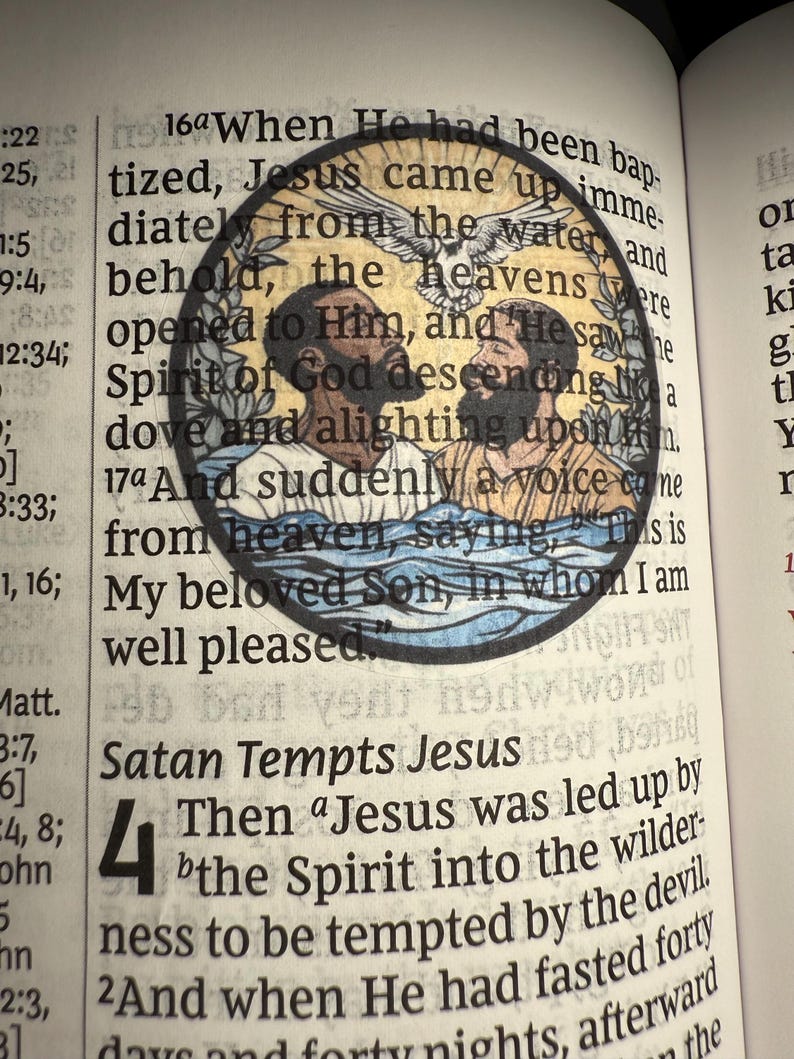 Puede incluir: Ilustraci&oacute;n circular de vidriera que representa una escena religiosa con dos figuras en el agua, una paloma y texto de la Biblia. La imagen est&aacute; situada sobre un libro abierto con texto impreso.