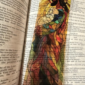 May include: A stained glass image of Jesus holding a lamb, set against a backdrop of open book pages. The artwork features vibrant colors and intricate details, with text from the Bible visible.