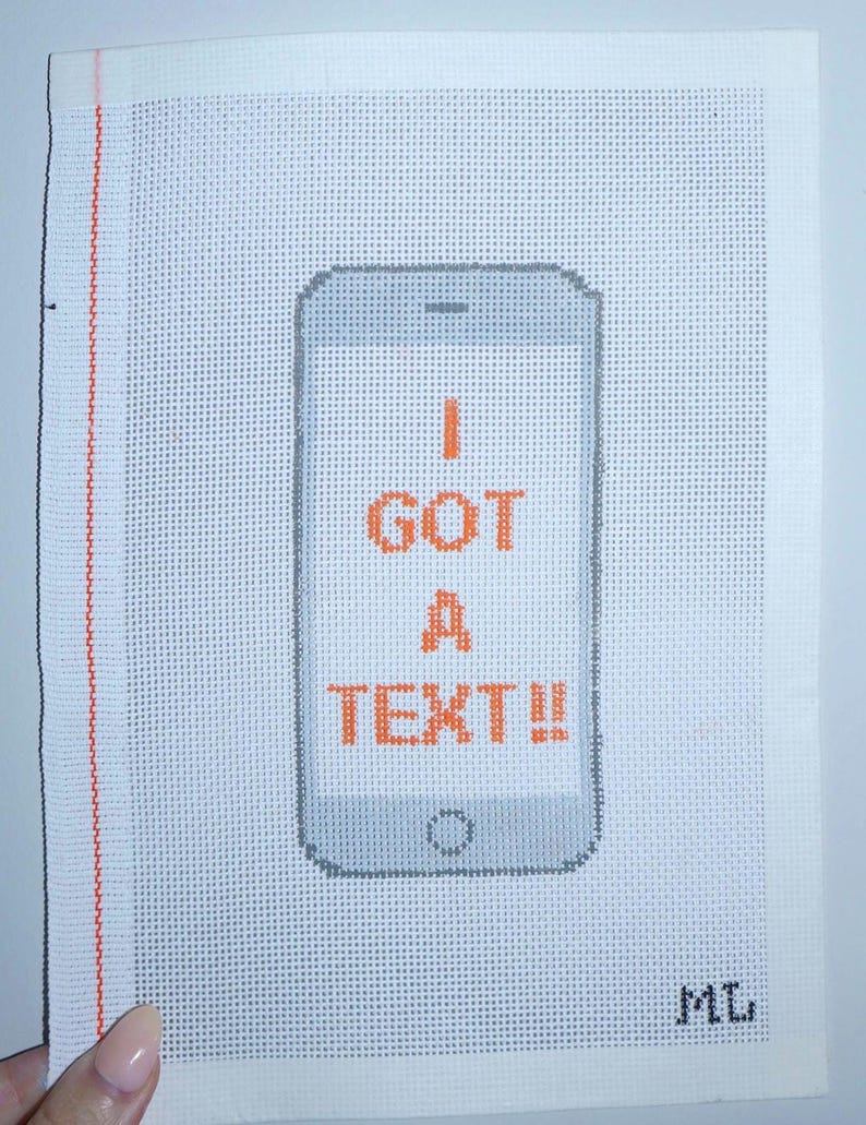 Puede incluir: Un lienzo blanco de punto de aguja con un dise&ntilde;o de tel&eacute;fono inteligente gris y el texto naranja "I GOT A TEXT!!". El lienzo tiene un borde vertical rojo en el lado izquierdo y las iniciales "ML" en la esquina inferior derecha.
