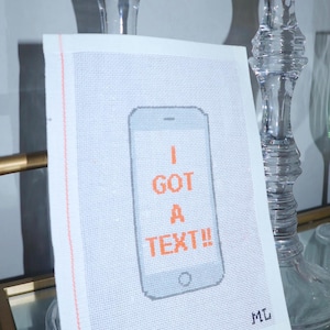 Puede incluir: Una pieza de punto de cruz blanca con un dise&ntilde;o de smartphone pixelado. El tel&eacute;fono es gris con texto naranja que dice "I GOT A TEXT!!". La pieza tiene un borde rosa y las iniciales "ML" en la esquina.