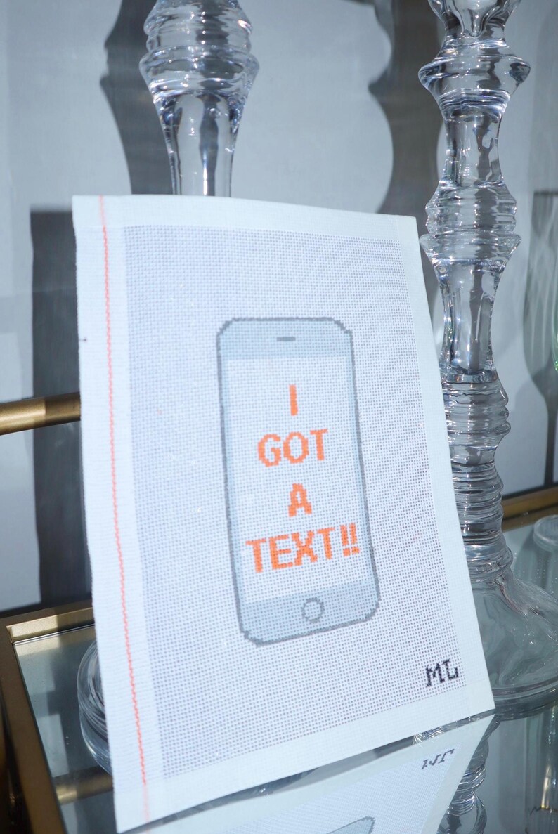 Puede incluir: Una pieza de punto de cruz blanca con un dise&ntilde;o de smartphone pixelado. El tel&eacute;fono es gris con texto naranja que dice "I GOT A TEXT!!". La pieza tiene un borde rosa y las iniciales "ML" en la esquina.