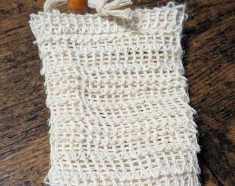 May include: A rectangular off-white soap pouch crafted from a textured, open-weave fabric. The top opening features a drawstring closure made of matching cord, secured with a single, smooth, round, polished brown wooden bead at the center.