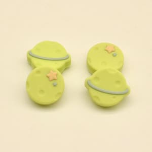 May include: Four light green planet-shaped embellishments with gray rings and star accents. The embellishments are round with a slightly textured surface. The background is a solid, light beige color. The text "ArtisanGlowCoCrafts" is at the bottom.