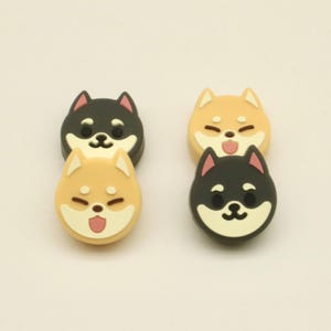 May include: Four Shiba Inu dog-shaped charms. Two are black and tan, and two are tan. Each charm has a smiling face with a pink tongue. The charms are arranged in two pairs, stacked on top of each other.
