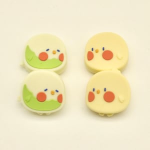 以下が含まれることがあります： 4つの鳥の形をした装飾品。2つはオレンジ色の頬と青い目をした薄い黄色です。他の2つは、緑の翼とオレンジ色の頬を持つ白です。すべて漫画のようなデザインで、白い背景に配置されています。