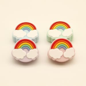 May include: Four colorful rainbow-shaped embellishments with white cloud accents. Each rainbow features a vibrant array of colors, including red, orange, yellow, green, blue, and purple. The embellishments are set against a light background.
