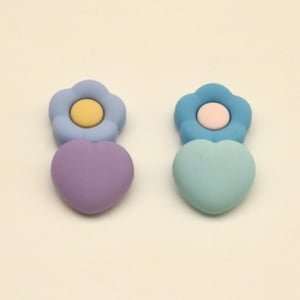 May include: Two decorative resin earrings. One earring features a light blue flower with a yellow center and a purple heart. The other earring has a light blue flower with a pink center and a light blue heart.