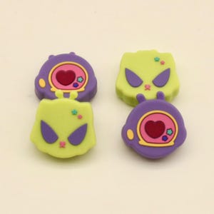 May include: Four colourful alien-themed charms. Two are purple with a pink heart and yellow accents, and two are lime green with purple eyes. The charms have a cartoonish design, likely for crafting or jewellery making.