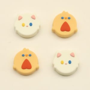 May include: Four colorful, cartoon-style embellishments. Two are white with pink ears, yellow and blue eyes. The other two are yellow with red beaks and blue eyes. The embellishments are in the shape of a cat and a chick.