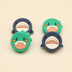 以下が含まれることがあります： 水色とネイビーブルーの漫画のサメ型装飾品4つ。各サメは、ギザギザの歯を持つ白い口、黒い目、オレンジ色のアクセントを持っています。装飾品は正方形に配置されています。