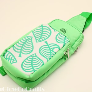 May include: A light green sling bag with a white panel featuring a repeating pattern of stylized green leaves. The bag has a zippered pocket, a green strap, and a small leaf logo. The bag is designed for carrying items.