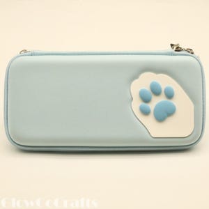 May include: A pale blue rectangular case with a white paw print and light blue paw pads. The case has a silver zip and is likely made of a soft material. The design is a cat paw.