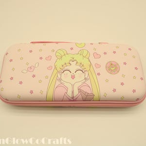 Puede incluir: Una funda rosa con una ilustración de dibujos animados de un personaje con cabello rubio y ropa rosa. La funda está decorada con estrellas, corazones y un emblema circular. Tiene una cremallera y un borde rosas.