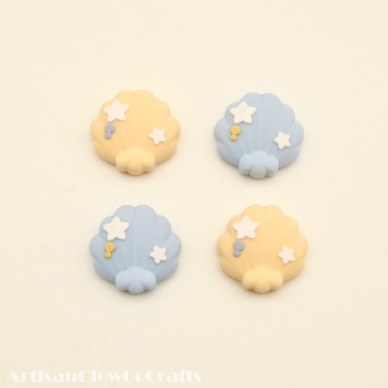 May include: Four seashell-shaped embellishments in pastel yellow and blue. Each shell is decorated with white stars and small yellow and blue accents. The embellishments are arranged on a light background.