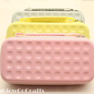 May include: Three small, rectangular cases in pastel shades: pink, yellow, and light green. Each case has a textured, dotted surface and a small handle. The cases are stacked on top of each other.