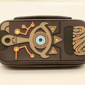 以下が含まれることがあります： 茶色の長方形ケースで、ハンドルが付いています。ベージュ、オレンジ、青色の詳細なデザインが特徴です。デザインには幾何学的な形状と、ゲームやファンタジーに関連するテーマを示唆する中央の目の様なエンブレムが含まれています。