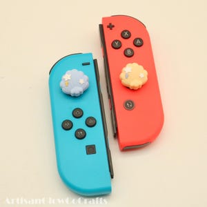 May include: Two Nintendo Switch Joy-Con controllers, one red and one blue. Each controller has a decorative cloud-shaped button. The blue controller has a light blue cloud with white stars, while the red controller has a yellow cloud.