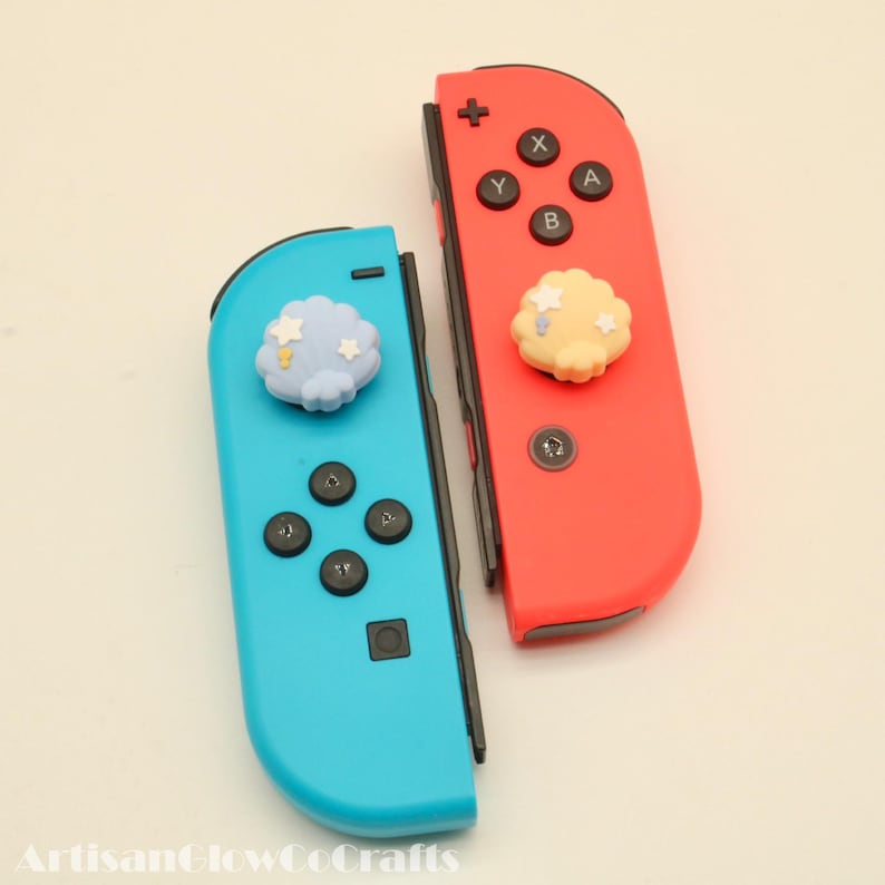 May include: Two Nintendo Switch Joy-Con controllers, one red and one blue. Each controller has a decorative cloud-shaped button. The blue controller has a light blue cloud with white stars, while the red controller has a yellow cloud.