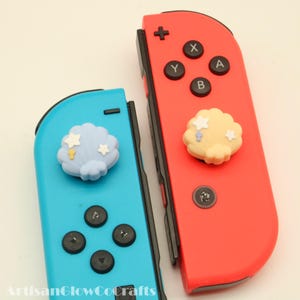 May include: A pair of Nintendo Switch Joy-Con controllers, one red and one blue. Each controller has a decorative cloud charm with star and raindrop accents. The controllers have black buttons and are set against a white background.