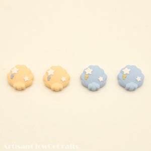 May include: Four cloud-shaped embellishments in soft yellow and blue. Each cloud features white star accents and a small yellow detail. The embellishments are arranged on a cream-colored background, perfect for craft projects.