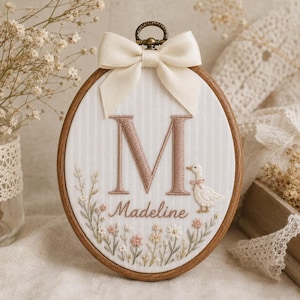 Op de afbeelding: Een ovale borduurring met witte stof met verticale strepen. De stof is voorzien van een grote geborduurde letter M in oudroze, de naam Madeline in cursief schrift, een kleine witte eend en een kleurrijk bloemstuk.