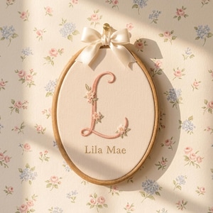 May include: An oval wooden embroidery hoop containing cream-colored fabric with a large, pink, floral-stitched letter L and the name Lila Mae embroidered in brown below it. A cream satin ribbon bow is attached to the top of the wooden frame.