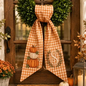 May include: A green boxwood wreath adorned with a long, orange and white gingham ribbon. The ribbon features an embroidered stack of three pumpkins on one side and an embroidered letter S surrounded by a leafy wreath on the other side.