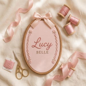 May include: An oval wooden embroidery hoop containing light pink fabric with the embroidered name Lucy BELLE in pink and brown thread, accented by floral vines. Includes pink ribbon, spools of pink thread, and gold-toned metal embroidery scissors.