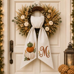 May include: A round grapevine wreath decorated with dried wheat, green foliage, and small white pumpkins. A long, cream-coloured fabric sash with thin vertical stripes hangs from the centre, featuring embroidered stacked pumpkins and the letter M.