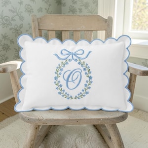 Peut inclure: Un coussin rectangulaire blanc avec un bord festonné garni de bleu clair. Le centre présente une lettre O brodée en bleu clair entourée d'une couronne florale de feuilles vertes et de fleurs bleues, surmontée d'un nœud bleu clair.