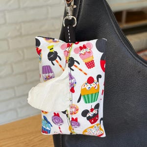May include: A white fabric tissue holder with a colorful print of cupcakes, Mickey Mouse ears, and other Disney characters. The holder is attached to a black leather purse with a silver clip.