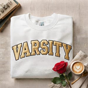 May include: A white crewneck sweatshirt featuring the word VARSITY in large, arched, gold-colored block letters with a black outline. The garment has a ribbed collar and long sleeves, presented in a clean, folded layout for a clear view of the design.