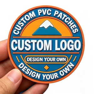 May include: A circular PVC patch featuring a mountain graphic with a white peak, orange and blue background, and white text reading CUSTOM PVC PATCHES, CUSTOM LOGO, and DESIGN YOUR OWN. The design includes orange and blue borders and a diamond shape.