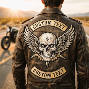 May include: A brown distressed leather jacket featuring a large back patch with a central skull graphic, wings, and two banners containing the text CUSTOM TEXT. The jacket has a classic collar and long sleeves, worn by an individual.