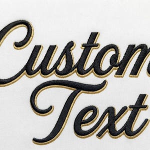 May include: The words Custom Text are embroidered in a black cursive script font with a gold outline. The lettering is positioned on a plain white fabric background. The embroidery features a raised texture and a clean, professional finish.