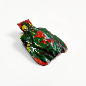 Könnte beinhalten: Eine grüne, rote und gelbe Spielzeug-Frosch aus Blech mit einem Jungen, der auf seinem Rücken reitet. Das Spielzeug ist aus Metall und hat ein einfaches Design. Der Junge trägt ein blaues Hemd und rote Shorts. Der Frosch hat einen gelben Bauch und schwarze Augen.