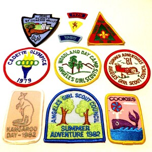 May include: A collection of vintage Girl Scout patches. The patches feature various designs, including a kangaroo, a torch, a whale, and text such as "Cadette Olympics 1979" and "Summer Adventure 1982". The patches are in various shapes and colors.