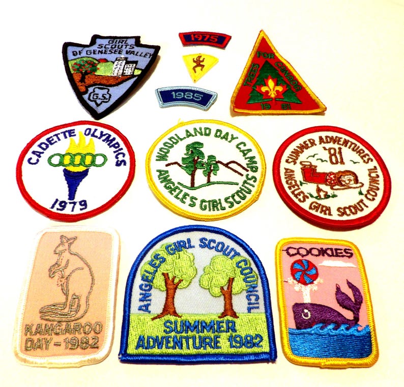 May include: A collection of vintage Girl Scout patches. The patches feature various designs, including a kangaroo, a torch, a whale, and text such as "Cadette Olympics 1979" and "Summer Adventure 1982". The patches are in various shapes and colors.