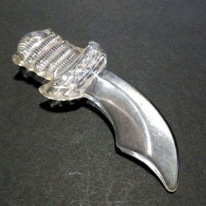 May include: A clear, curved glass object with a textured top and a smooth, polished handle. The top section has a ribbed design with small, embedded embellishments. The object is set against a dark background, highlighting its transparent nature.